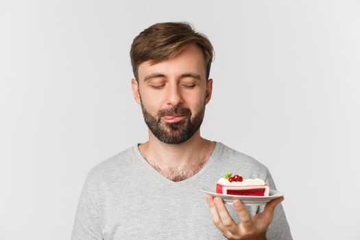 Close-up of handsome bearded man, smiling and looking at tasty cake, standing over white background.