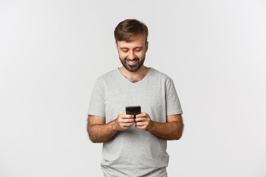 Portrait of handsome modern guy with beard, messaging and looking at smartphone screen, standing over white background.