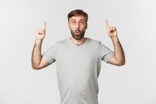 Portrait of adult caucasian guy with beard, wearing gray t-shirt, looking impressed at something cool, pointing fingers up, showing promo, standing over white background.