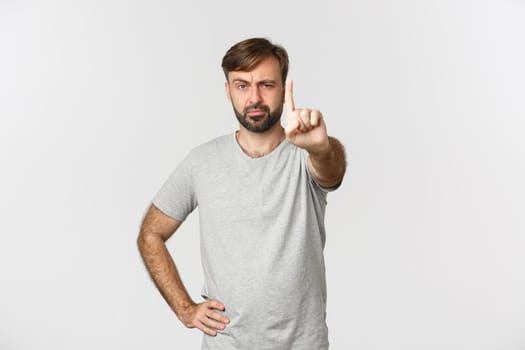 Portrait of serious adult man with beard, wearing gray t-shrit, shaking finger with disappointed face, scolding someone and prohibit action, standing over white background.