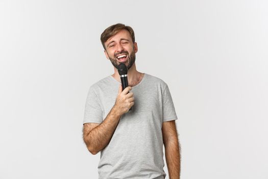 Handsome carefree guy singing song in karaoke, holding microphone, standing over white background.