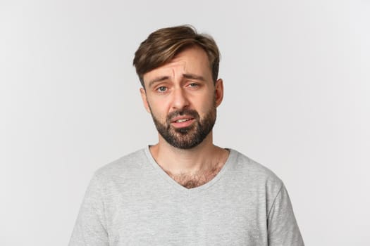 Skeptical and unamused bearded man in gray t-shirt, frowning and looking exhausted, standing over white background.
