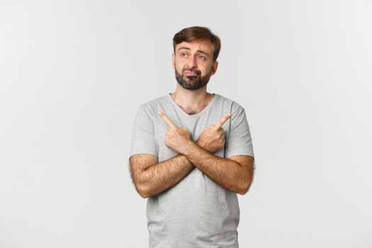 Portrait of indecisive beaded man in gray t-shirt, making decision, pointing fingers sideways at two choices, standing over white background.