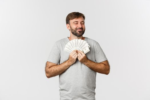 Image of handsome bearded guy who loves money, hugging cash and smiling satisfied, standing over white background.