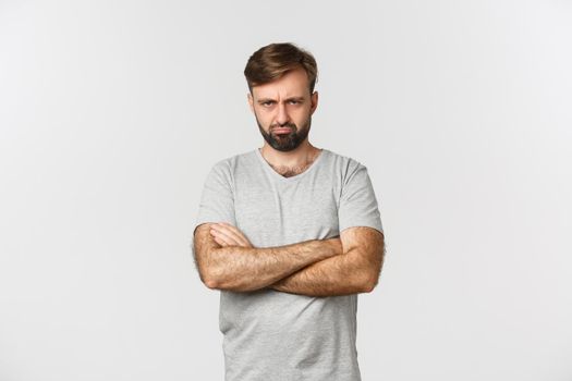 Portrait of offended handsome man, sulking and making angry grimace, cross hands over chest defensive, standing over white background.