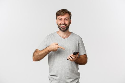 Portrait of excited smiling man in gray t-shirt, pointing at mobile phone, showing application, standing over white background.