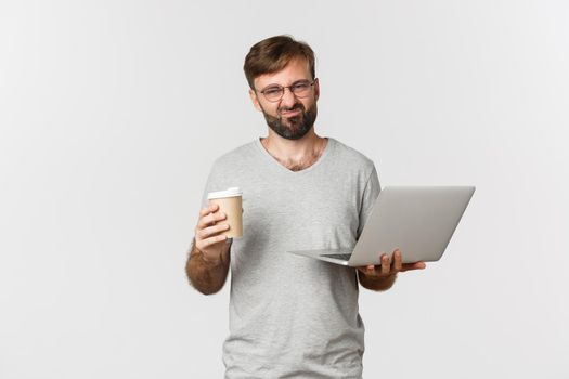 Image of handsome bearded man, holding laptop and drinking bad coffee, grimacing from dislike, standing over white background.