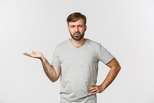 Portrait of confused and annoyed guy arguing, raising hand up and looking puzzled, standing over white background.