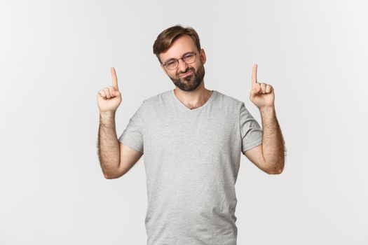 Skeptical and unamused bearded man smirking, pointing fingers up at something bad, showing logo, standing over white background.