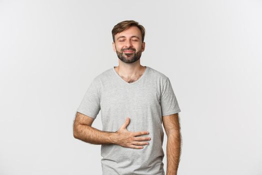 Portrait of happy man feeling pleased after eating tasty food, rubbing belly and smiling delighted, standing over white background.