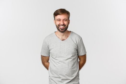 Image of happy bearded man in gray t-shirt, smiling and laughing, standing humble with hands behind.