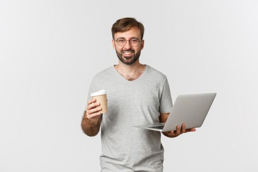 Portrait of handsome male freelancer, drinking coffee and working with laptop, standing over white background.