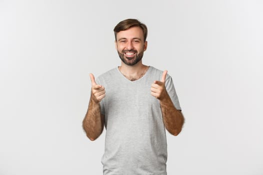 Image of satisfied smiling man in gray t-shirt, showing thumbs-up in approval, like something good, white background.