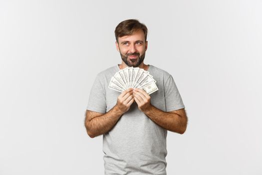 Greedy smiling man with beard, thinking about shopping, holding money and looking at upper left corner, standing over white background.