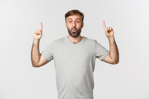 Image of excited handsome man in gray t-shirt, pointing fingers up and saying wow, standing over white background.