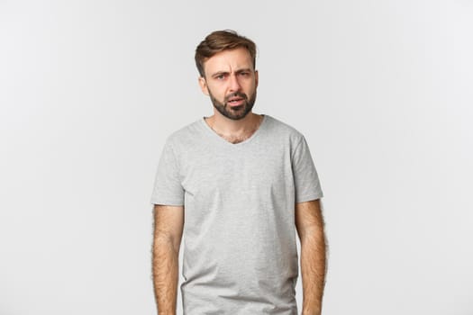 Image of confused bearded man in casual t-shirt, looking puzzled at camera and frowning, dont understand something, standing over white background.