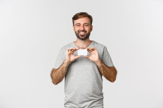 Cheerful caucasian man in gray t-shirt, showing credit card and smiling, excited for shopping, standing over white background.