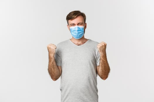 Concept of pandemic, coronavirus and social-distancing. Cheerful caucasian man in gray t-shirt and medical mask, winning something, rejoicing from win, standing over white background.