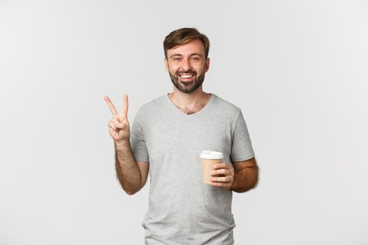 Image of happy guy drinking takeaway coffee, showing peace sign and smiling, standing over white background.