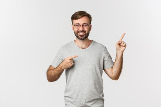 Cheerful bearded man smiling, pointing fingers at upper right corner, showing logo, standing over white background.