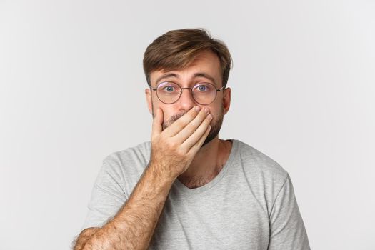 Close-up of shocked and worried man in glasses, gasping and cover mouth, hear something scary, standing over white background.