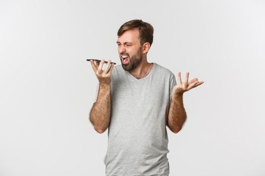 Portrait of angry man in gray t-shirt, having argument on speakerphone, shouting at mobile phone with mad face, standing over white background.