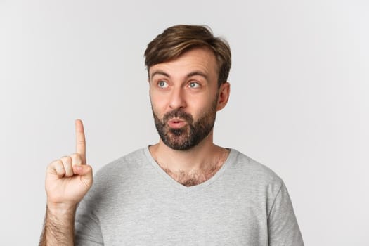 Close-up of thoughtful bearded man in gray t-shirt, having idea, raising finger and looking up, standing over white background.