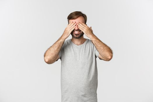 Image of happy smiling guy with beard, close eyes and waiting for surprise, anticipating something, standing over white background.