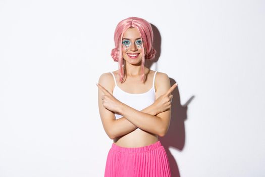 Smiling confident young woman in party costume and pink wig looking satisfied, pointing fingers sideways, showing left and right choices, standing over white background.