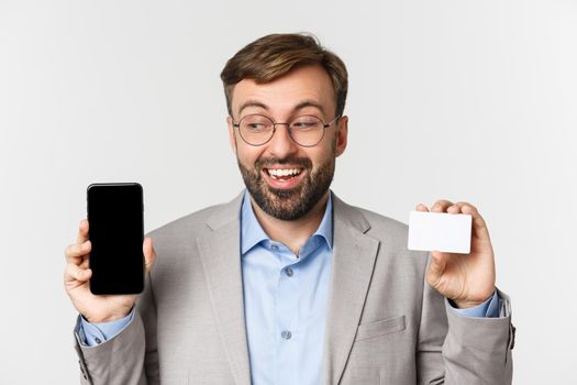 Close-up of handsome smiling male entrepreneur, wearing glasses and gray suit, showing credit card and looking excited at smartphone screen, standing over white background.