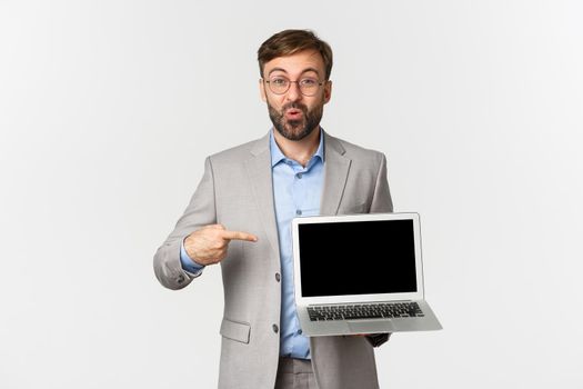 Image of smiling handsome businessman in grey suit and glasses, pointing finger at laptop screen, showing presentation or advertisement, standing over white background.