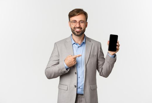 Handsome smiling businessman with beard, wearing grey suit and glasses, pointing finger at mobile phone screen, showing app, standing over white background.