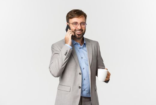 Image of successful businessman in grey suit and glasses, drinking coffee and talking on phone with pleased smile, standing over white background.