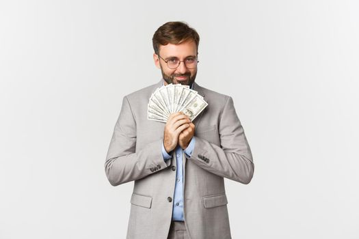 Image of happy businessman with beard, wearing grey suit and glasses, holding money and thinking about shopping, smiling greedy, standing over white background.