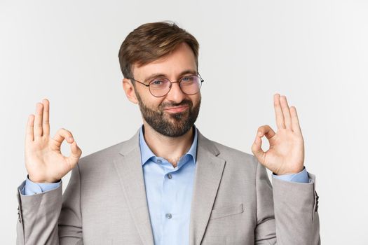 Close-up of impressed boss in gray suit and glasses, showing okay sign and smiling, give approval, praise good job, standing over white background.