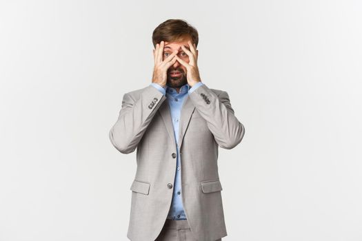 Portrait of ambushed businessman in grey suit, cover eyes and peeking through fingers with scared face, standing over white background.