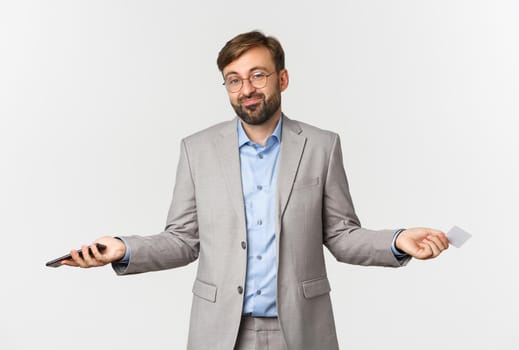 Image of clueless smiling businessman in glasses and gray suit, spread hands sideways and shrugging confused, holding mobile phone and credit card.