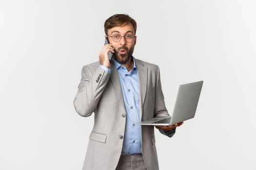 Portrait of handsome businessman in grey suit and glasses, receive good news during phone call, holding laptop, looking satisfied, standing over white background.