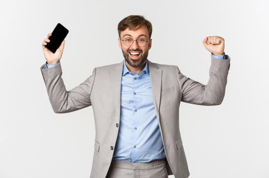 Image of successful businessman in grey suit and glasses, holding mobile phone, raising hands up and shouting yes from satisfaction, triumphing over white background.