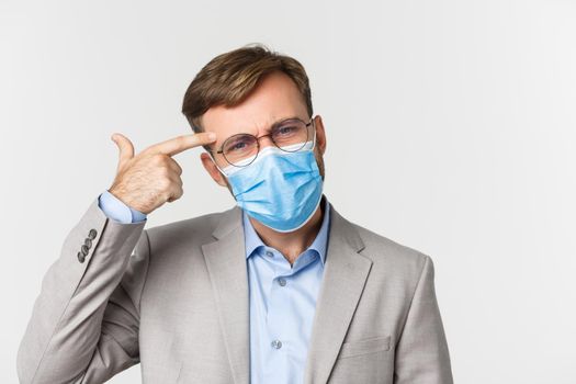 Concept of work, covid-19 and social distancing. Close-up of angry boss scolding person for acting stupid, pointing at head and frowning, wearing medical mask during pandemic, white background.