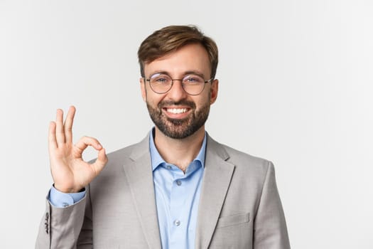 Close-up of handsome confident businessman, wearing glasses and gray shirt, showing okay sign and smiling, approve something, standing over white background.