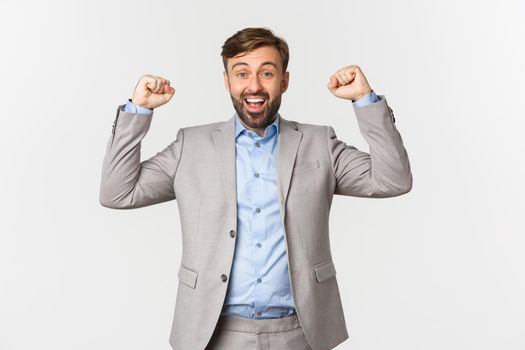 Portrait of relieved and happy bearded man in business suit, rejoicing over win, raising hands up and saying yes with satisfaction, standing over white background.