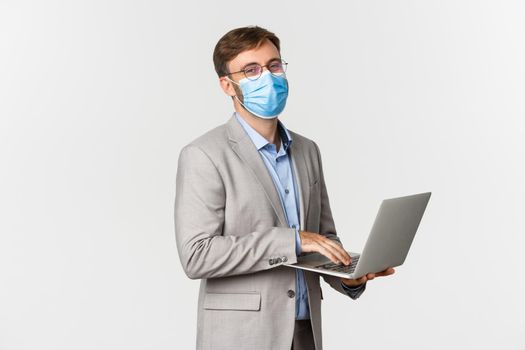 Concept of work, covid-19 and social distancing. Image of working businessman in medical mask and suit, using laptop and smiling happy, standing over white background.