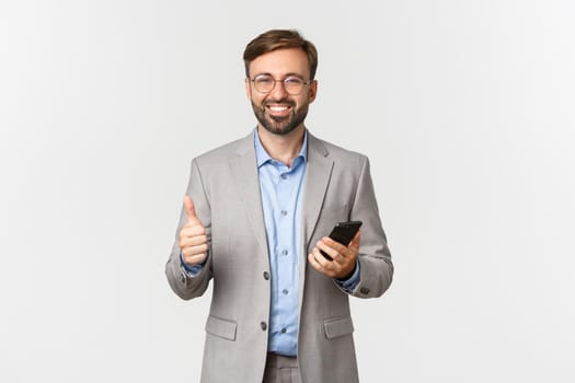 Portrait of handsome successful businessman with beard, wearing glasses and gray suit, holding mobile phone and showing thumbs-up in approval, smiling pleased, white background.