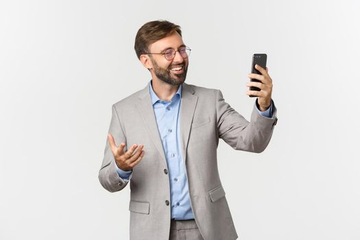 Portrait of handsome smiling businessman with beard, wearing grey suit and glasses, having video call with business partners, using mobile phone, standing over white background.