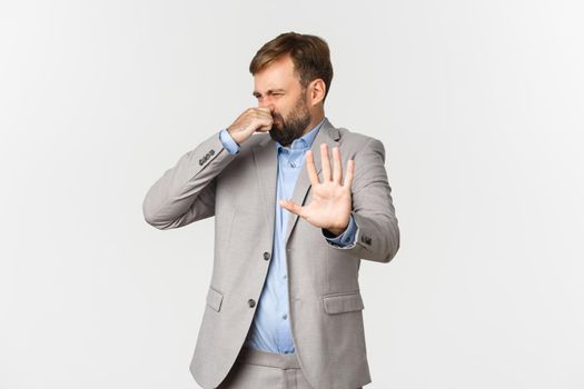 Portrait of disgusted bearded businessman in grey suit, rejecting something with awful smell, shut nose and making decline sign, standing over white background.
