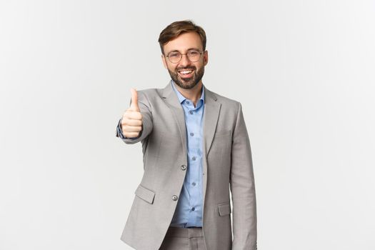 Portrait of handsome smiling businessman with beard, wearing glasses and grey suit, showing thumbs-up satisfied, praising good work, standing over white background.