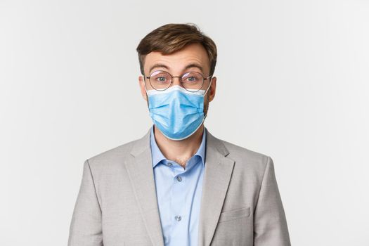 Concept of work, covid-19 and social distancing. Close-up of surprised man in gray suit and medical mask, raising eyebrows and looking amazed at camera, standing over white background.
