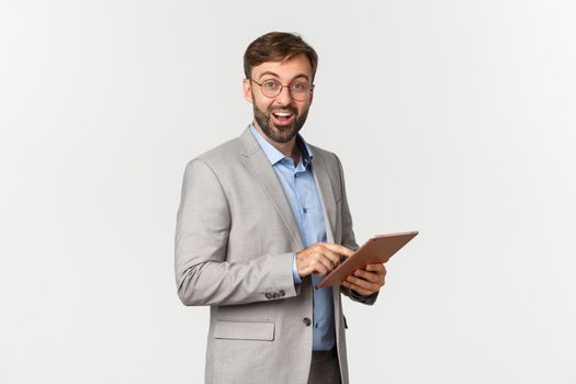 Portrait of happy and surprised bearded businessman in grey suit and glasses, holding digital tablet, smiling amazed at camera, standing over white background.