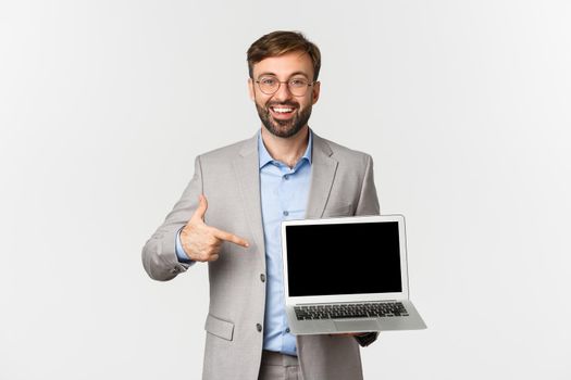 Image of smiling handsome businessman in grey suit and glasses, pointing finger at laptop screen, showing presentation or advertisement, standing over white background.